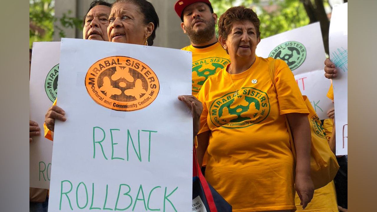 A woman, left, holds up a sign that says "Rent Rollback," while standing next to people wearing yellow shirts.
