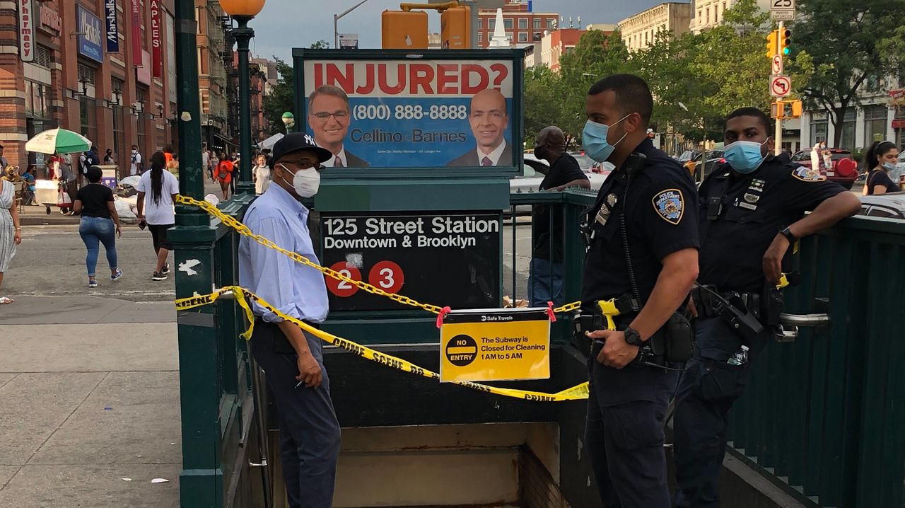 Outside 125th Street Station, yellow tape runs across the entrance. An NYPD officer dressed in black, stands a few inches in front of the left banister, while another man, wearing black pants and a light blue dress shirt, stands about six inches to the left of the right banister. Another man, wearing a light blue face mask and a black suit jacket and pants, stands on the opposite end of the right banister.