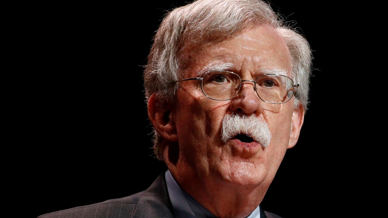 Former national security adviser John Bolton, wearing a dark brown suit jacket, a sky blue dress shirt, and a square glasses, stands against a black background.