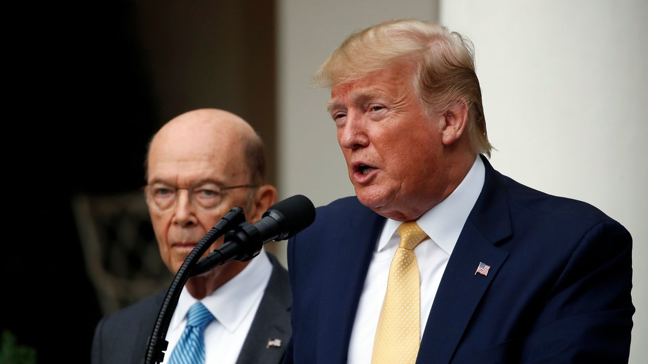 President Donald Trump, right, wearing a navy blue suit jacket, a yellow tie, and a white dress shirt, speaks into a black microphone. A few feet to his right stands a man, wearing circular glasses, a white dress shirt, a teal tie with white stripes, and a black suit jacket.