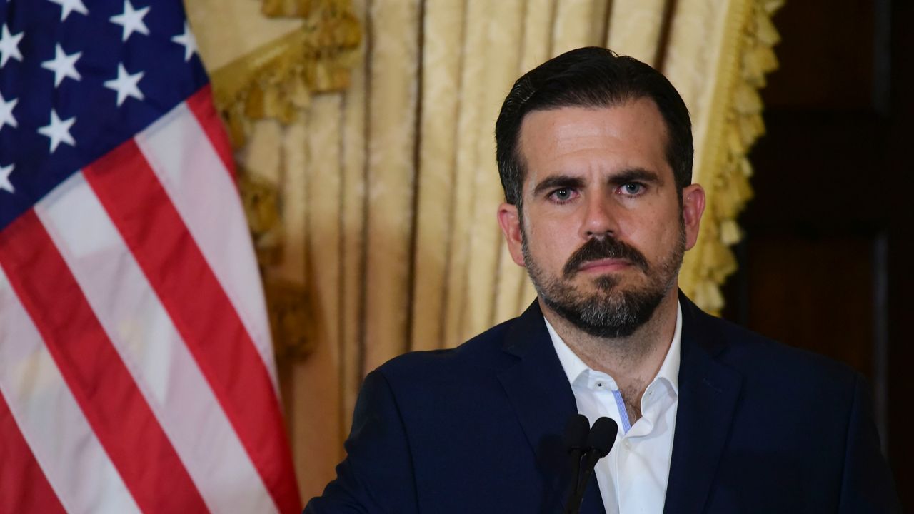 A man, wearing a white dress shirt and a dark navy blue blazer, stands behind a black microphone. He stands in front of a red, white, and blue American flag with white stars, and ruffled yellow curtains