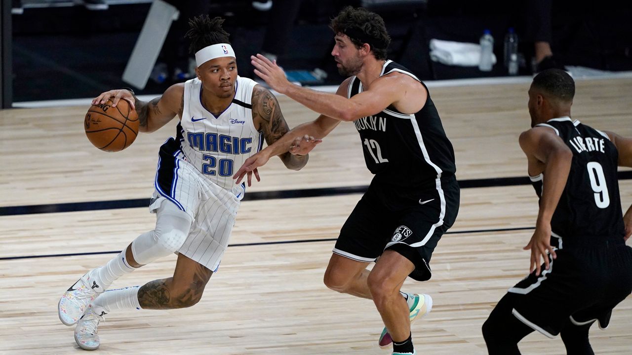 Markelle Fultz, left, in a white Orlando Magic uniform, driving with a basketball in his right hand, up against a Brooklyn Nets player, center, wearing a black uniform.