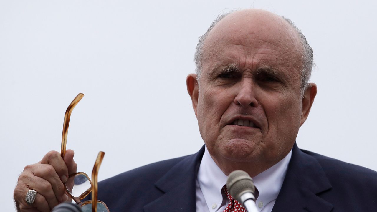 Rudy Giuliani, wearing a black suit jacket, a white dress shirt, and a red-and-black- tie, holds square glasses with light brown plastic frames while speaking into a silver microphone and standing in front of a solid white background. He wears a silver, square ring on his right pinky finger.