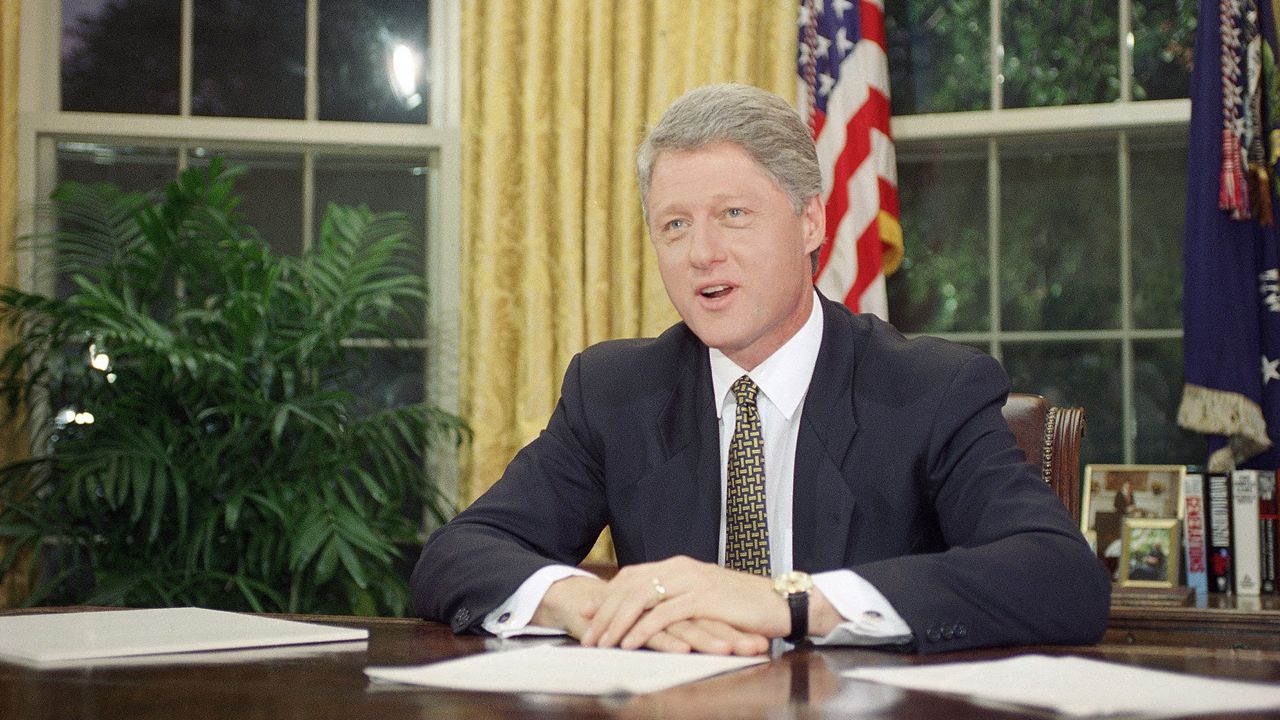 Bill Clinton, wearing a black suit jacket, a white dress shirt, an olive green tie, and a watch with a black band, sits against the brown wooden Resolute Desk, upon which three stacks of white paper sit.