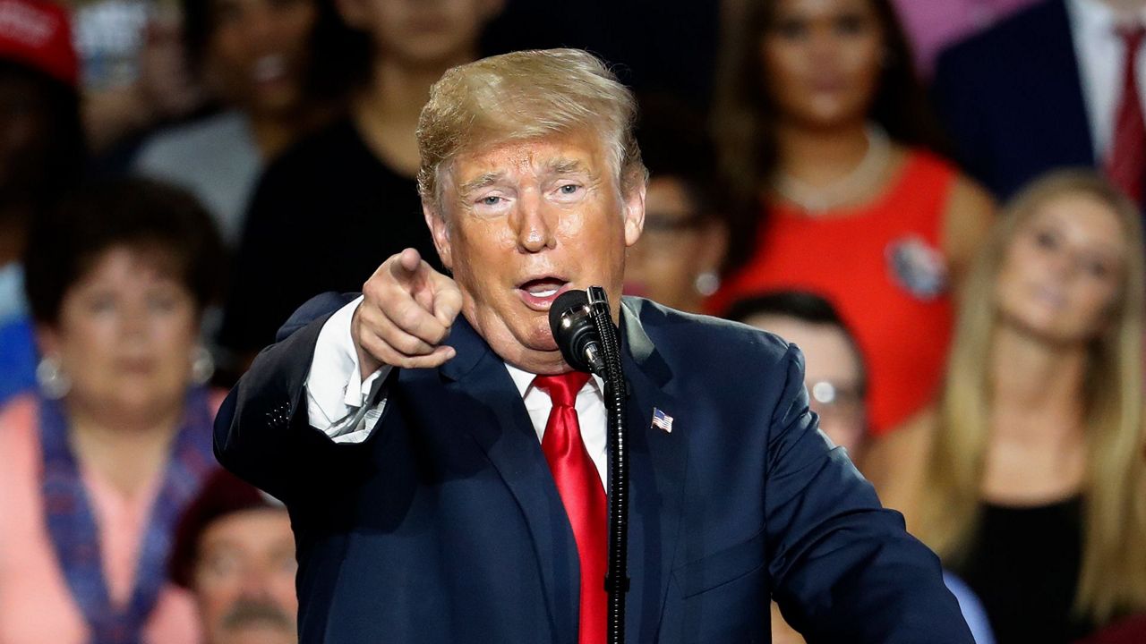 Donald Trump, wearing a navy blue suit jacket, a white dress shirt, and a red tie, points near a blue lectern. A crowd of people are behind him.