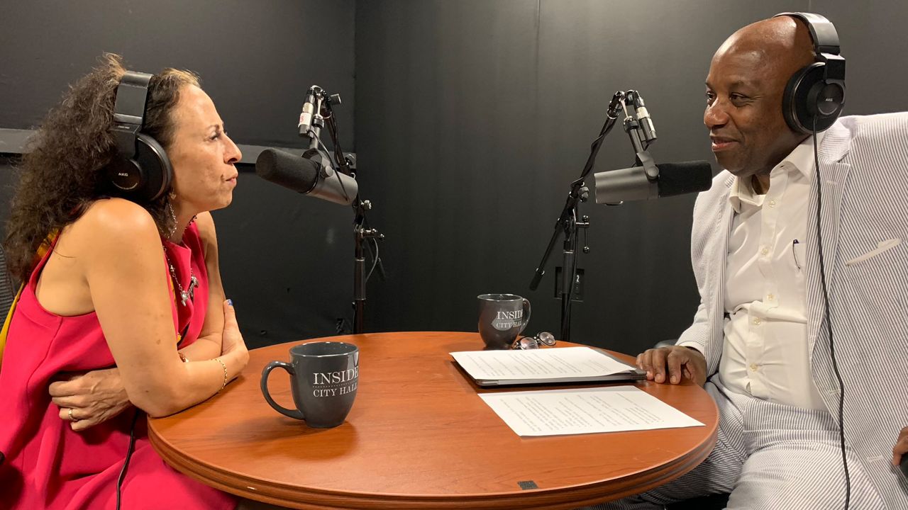 A woman, left, wearing a pink dress and black headphones, looks into a black microphone while sitting at a circular brown table. Two black mugs with white text that read "Inside City Hall" sit on the table, near sheets of white paper. "You Decide" Host Errol Louis, wearing a white dress shirt and a white suit, wears black headphones and looks into a black microphone.