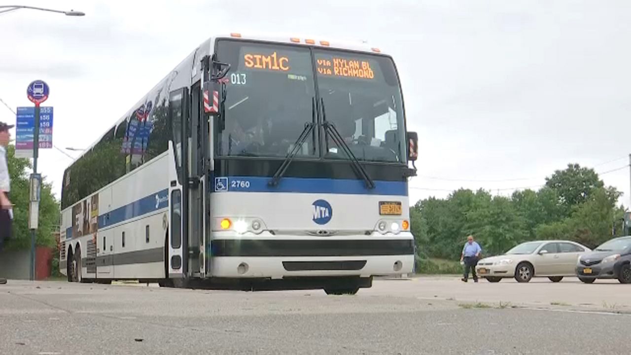 A bus parked on a sidewalk. Orange electronic text reads "SIM1C via Hylan Bl via Richmond" on the top of a bus.