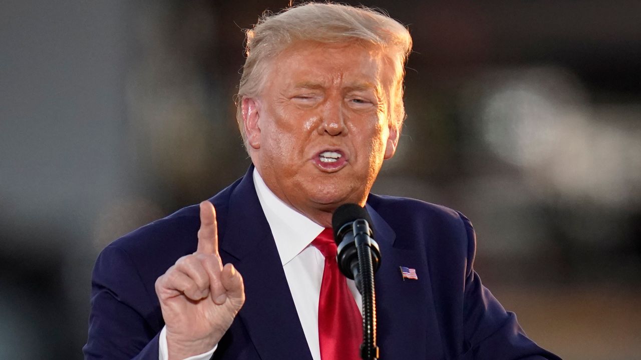 President Donald Trump, wearing a navy blue suit jacket with a red-white-and-blue American flag pin placed on the left side of his chest, a white dress shirt, and a red tie, speaks into a black microphone.