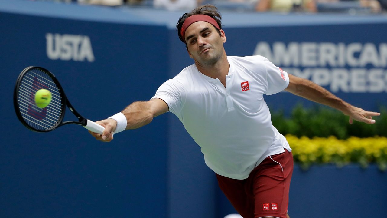 Roger Federer, wearing a white polo shirt, red shorts, a white wristband, and a red headband, reaches out with a black racket with a white handle. A green tennis ball makes contact with the center of the racket.