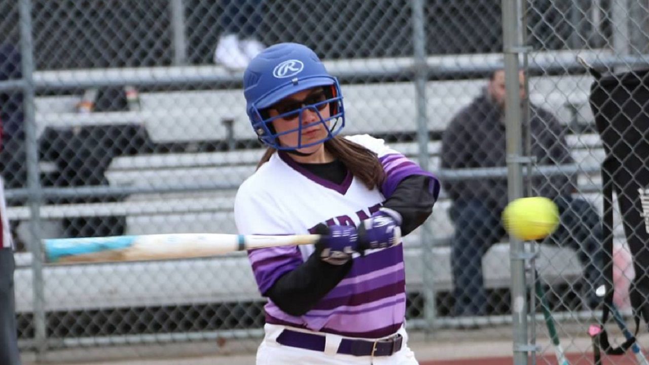 NY1 Scholar Athlete Megan Poser, wearing a royal blue softball helmet with a face guard, a white and purple jersey, and swinging a bat at a yellow tennis ball.