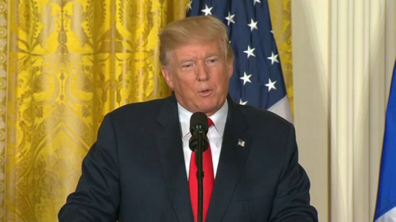 President Donald Trump in a black suit, a white shirt, and a red tie. He stands in front of a microphone. Yellow and white tapestry and an American flag are behind him.