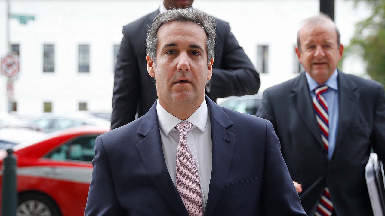 Michael Cohen, center, wearing a navy blue suit jacket, a white dress shirt, and a light pink tie. Two people wearing suits stand behind him.