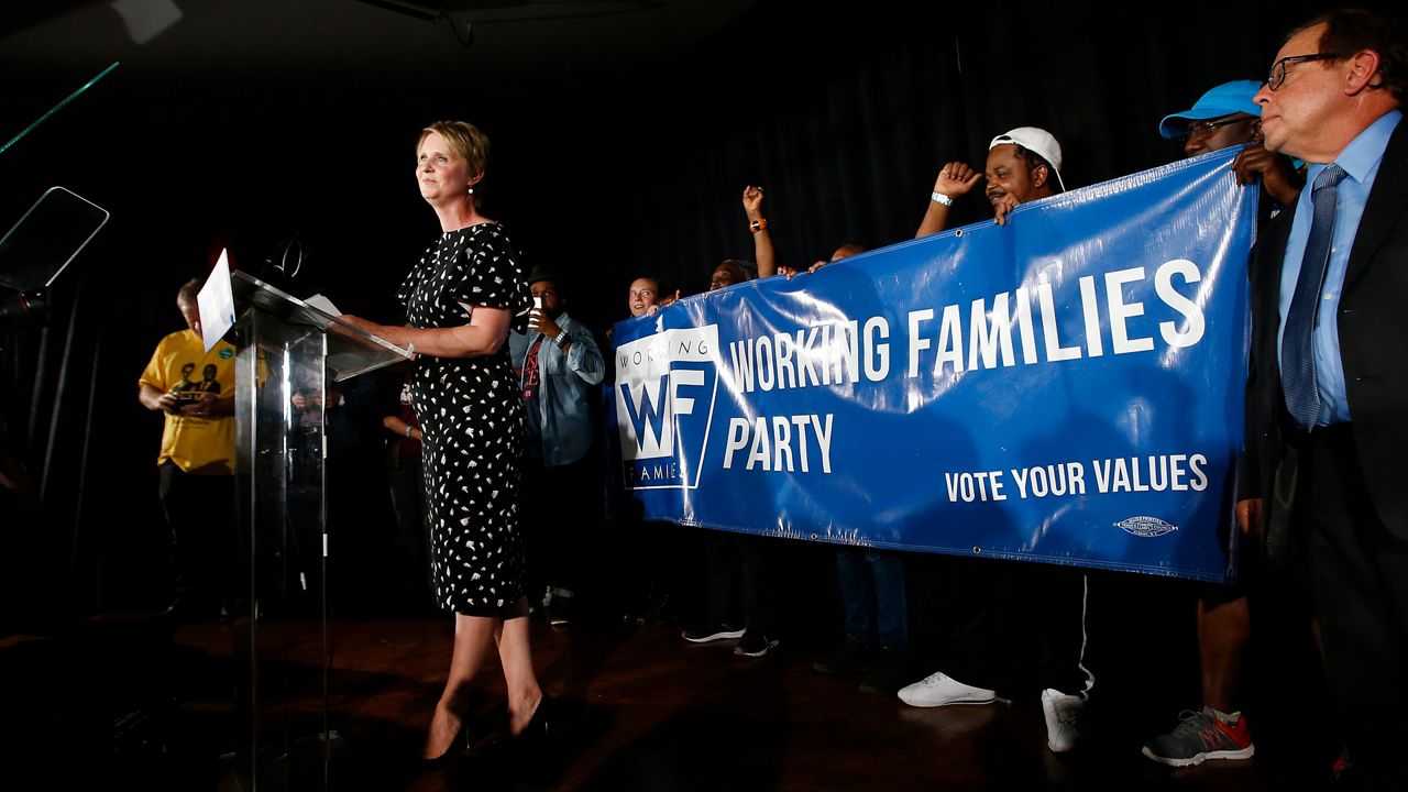 A woman, left, wearing a black dress with white spots, stands at a clear glass podium. Behind her on a stage, people hold up a blue banner with white text that reads "Working Families Party Vote Your Values"