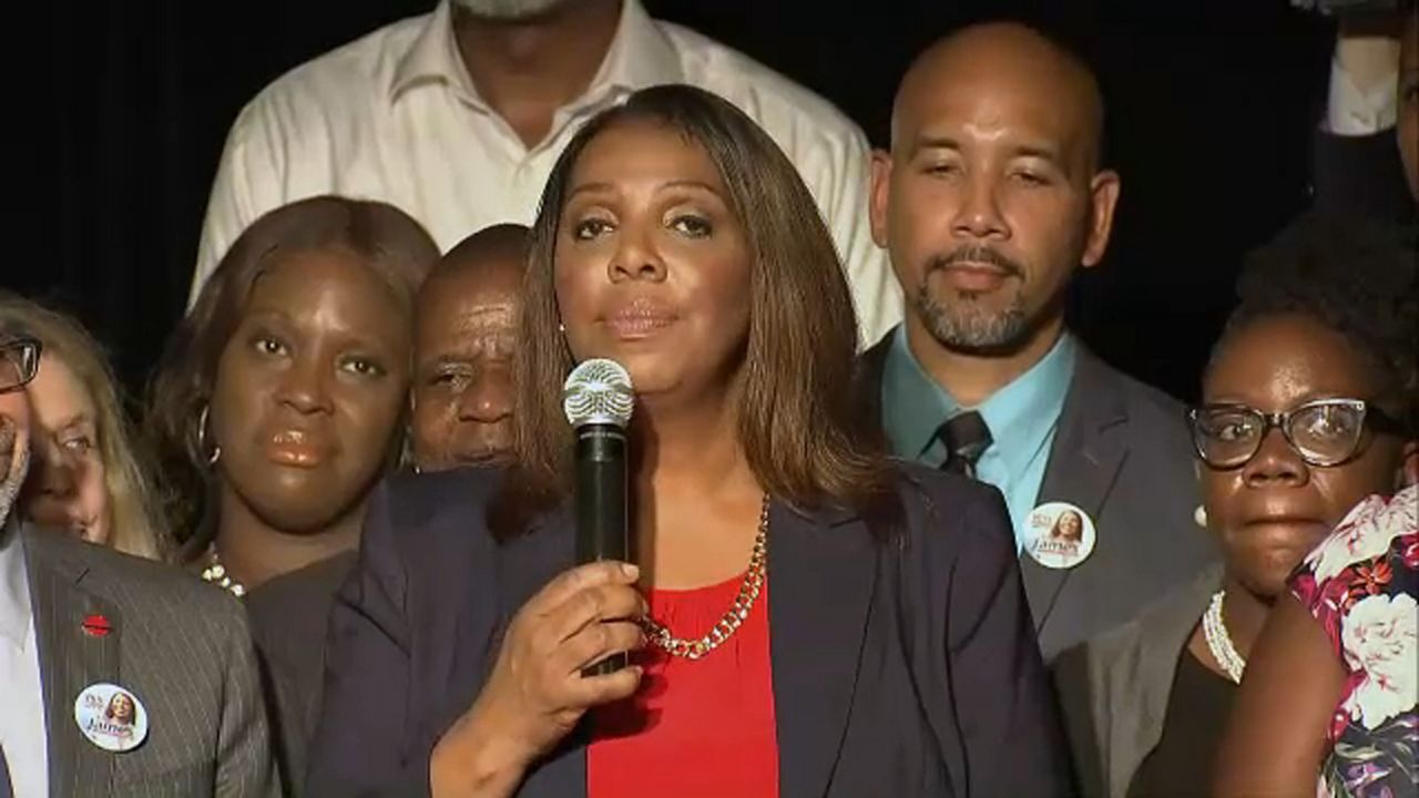 A woman, center, wearing a black suit jacket and a red shirt, holds a black microphone in her right hand. A group of people stand behind her.