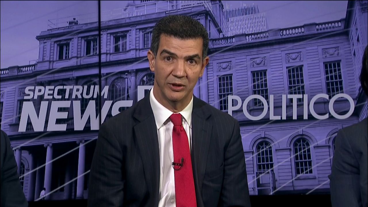 A man, center, wearing a black suit jacket, a white dress shirt, and a red tie, sits in front of a purple monitor displaying an image of City Hall. White text shows the logos for Spectrum News NY1 and Politico.