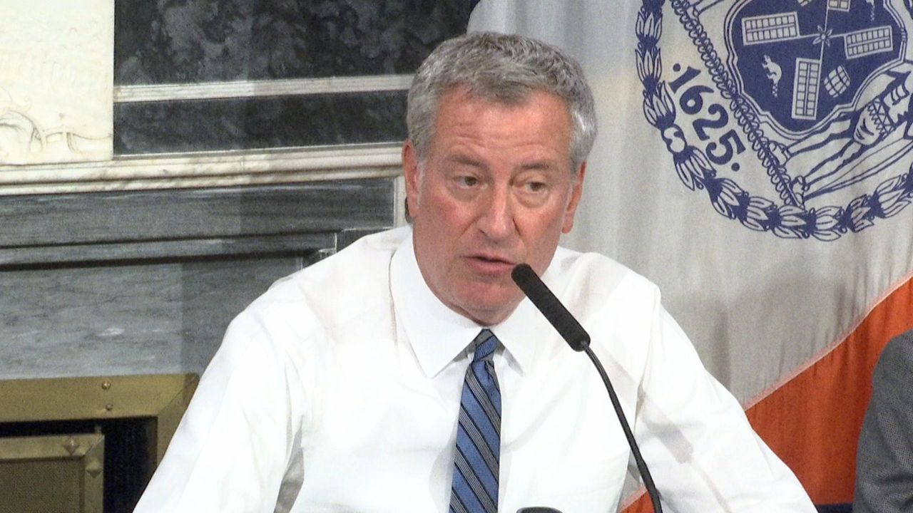 New York City Mayor Bill de Blasio, wearing a white dress shirt and a grey tie with black stripes, speaks into a long black microphone.