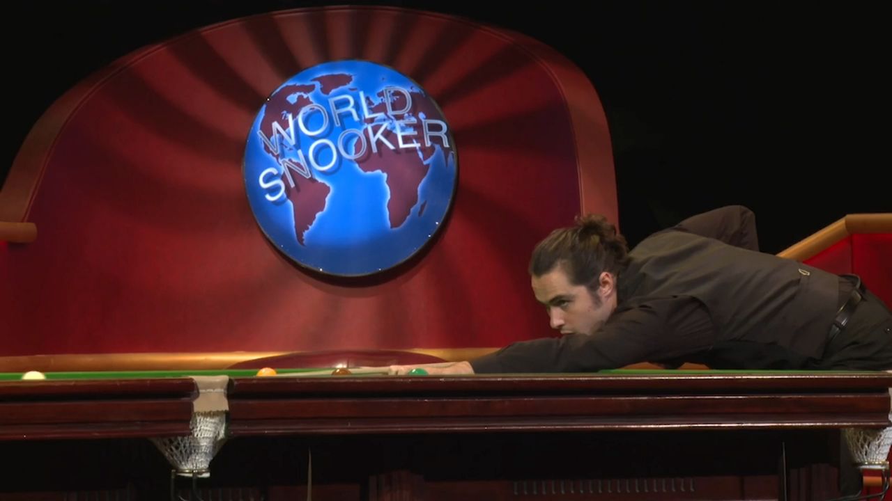 A man, right, leaning down on a table while holding a long pool stick. A sign featuring a globe reads "World Snooker" behind the man.