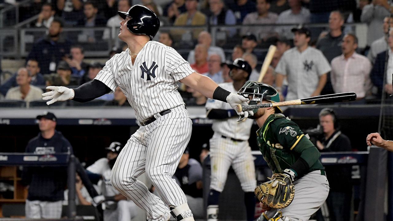Luke Voit, left, wearing a Yankees uniform while swinging back a brown-and-black bat near a catcher in green.