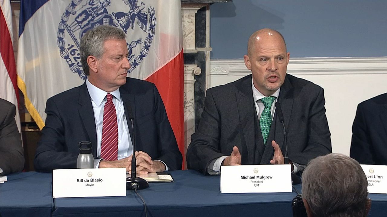 New York City Mayor Bill de Blasio, left, wearing a black suit jacket, a white dress shirt, and a red tie with white stripes. A man, right, wears a black suit jacket, a black vest, a white dress shirt, and a green tie with white spots. Both men sit at a table with a blue tablecloth, with black microphones and white placards in front of them. A blue, white, and orange New York City stands about five feet behind de Blasio, in front of marble and a blue-and-white wall.