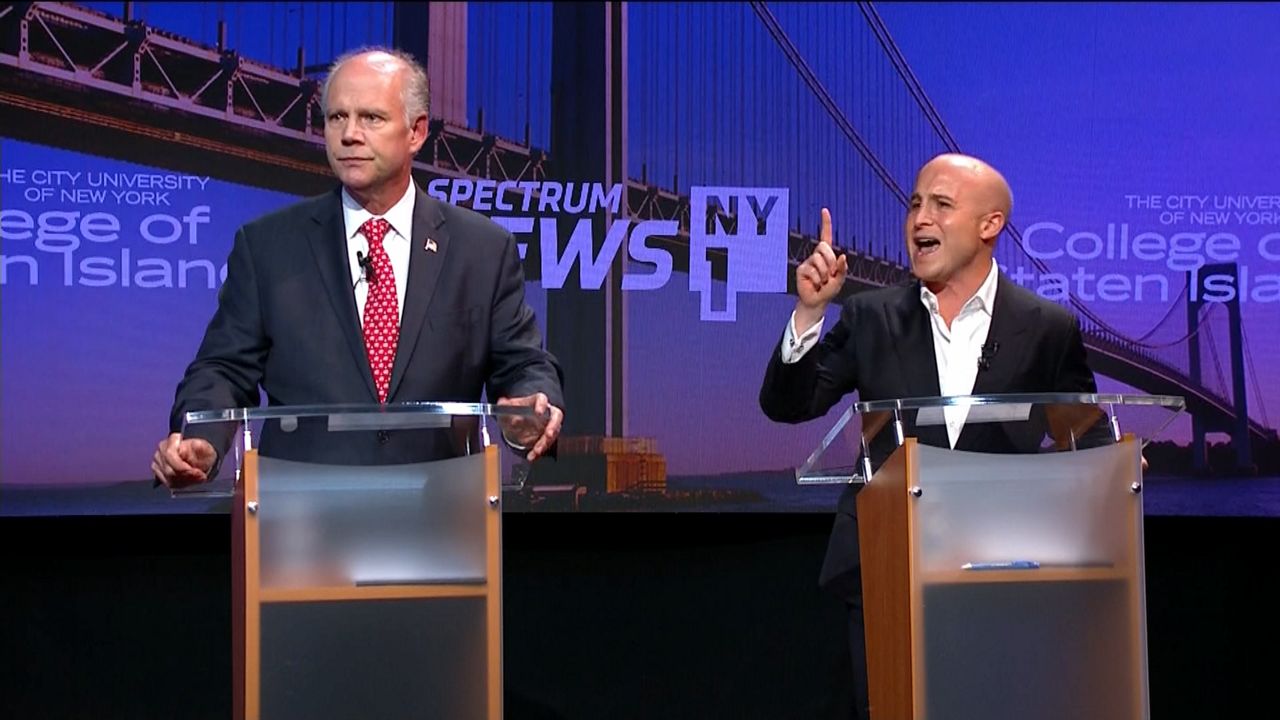 From left to right against a Spectrum News NY1 and College of Staten Island purple background: A man, left, wearing a black suit jacket, a white dress shirt, and a red tie with white spots; a man, right, wearing a white dress shirt and a black blazer. Both men stand in front of brown podiums.