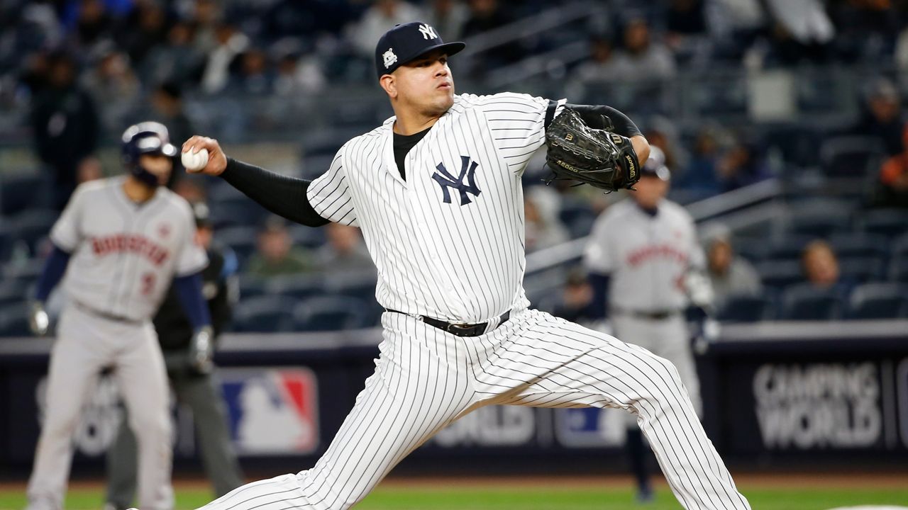 Dellin Betances, wearing a white New York Yankees pinstriped jersey, a black baseball cap with a white Yankees logo, a black sleeve on his right arm, and a black baseball glove on his left hand, holds a white baseball in his right hand as he enters his pitching motion.
