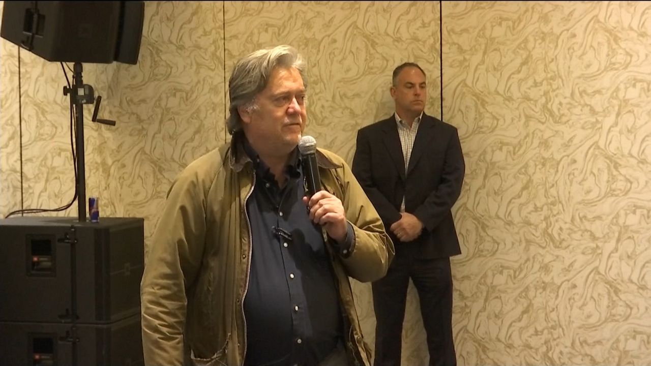 Steve Bannon, foreground, wearing a black dress shirt and a beige jacket while holding a black microphone.