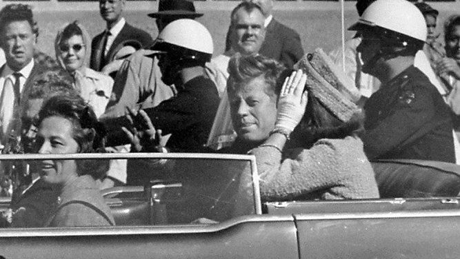 John F. Kennedy and Jackie Onassis wave in an open-top limo. 