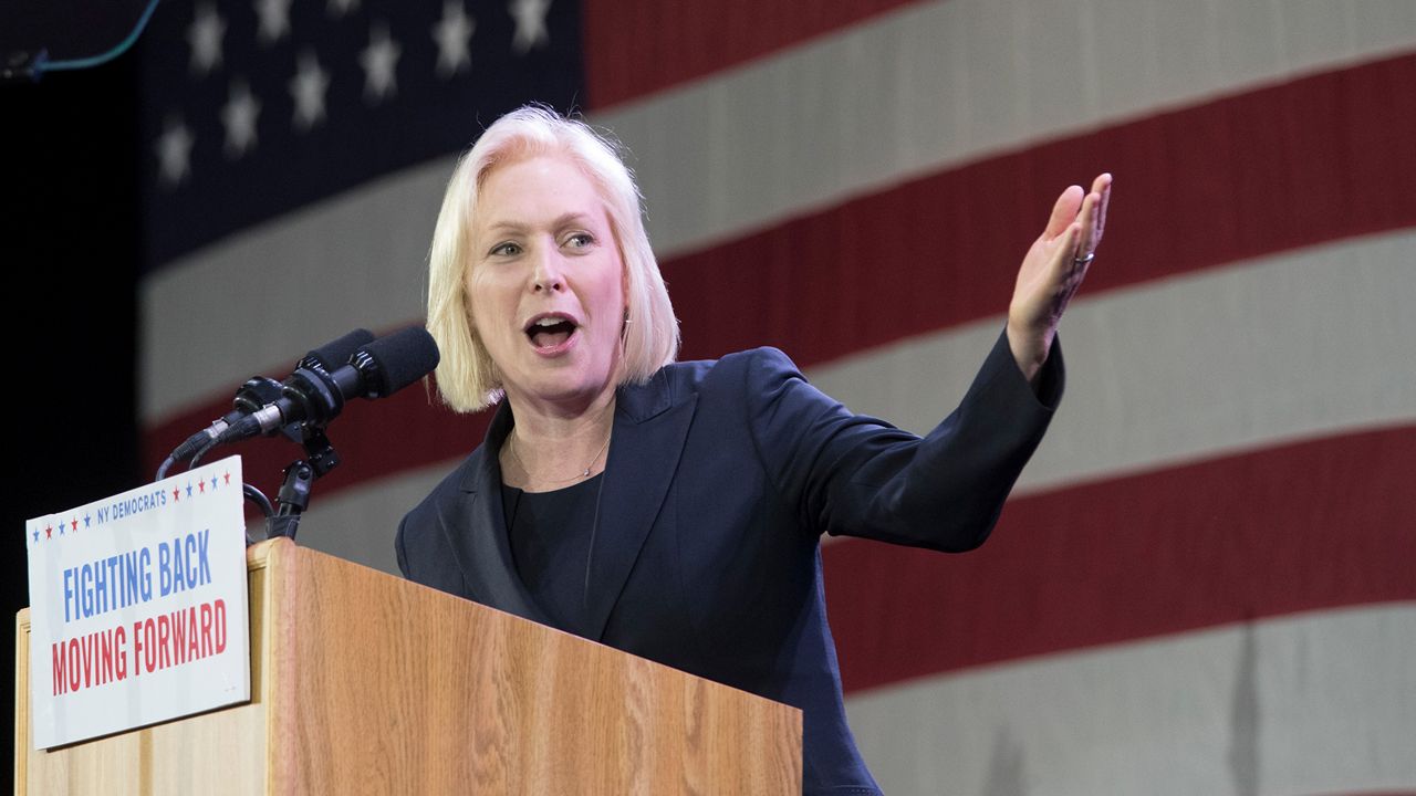 New York Sen. Kirsten Gillibrand, wearing a black shirt and a black blazer, stands at a brown lectern in front of a giant American flag. She speaks into a two black microphones connected by black wires jutting out from behind a white poster. Blue and red text, respectively, on the card read "Fighting Back Moving Forward"