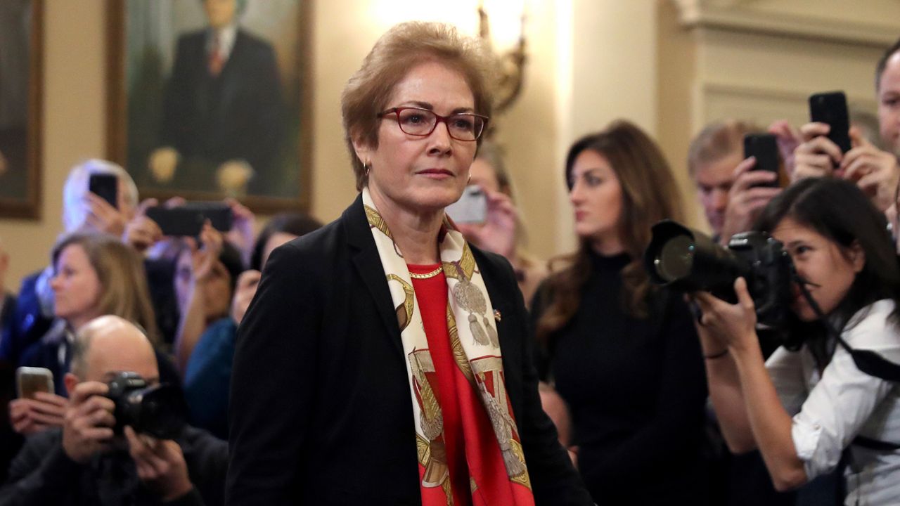 A woman, wearing square glasses, a black suit jacket, a red blouse, and a light yellow scarf, stands amid a crowd of people, some of whom hold black cameras to their faces.