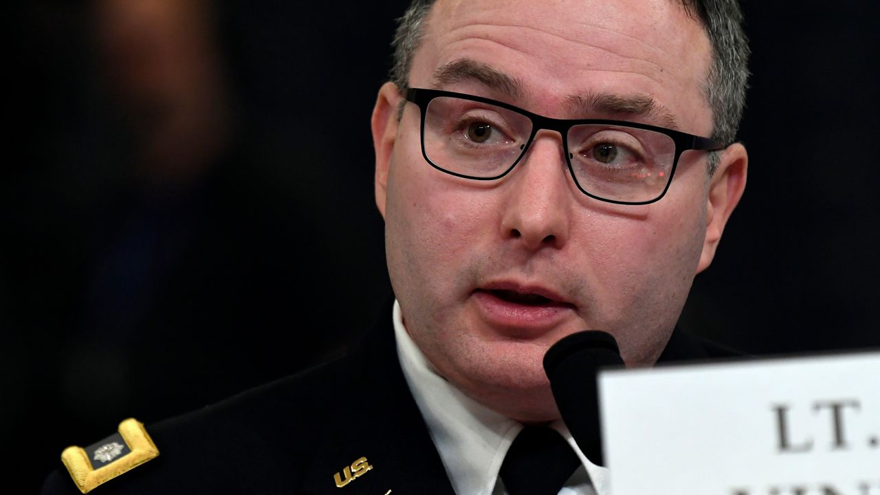 A man, wearing black, square-framed glasses, a white dress shirt, a black tie, and a black military uniform, speaks into a black microphone behind a white placard with black text.