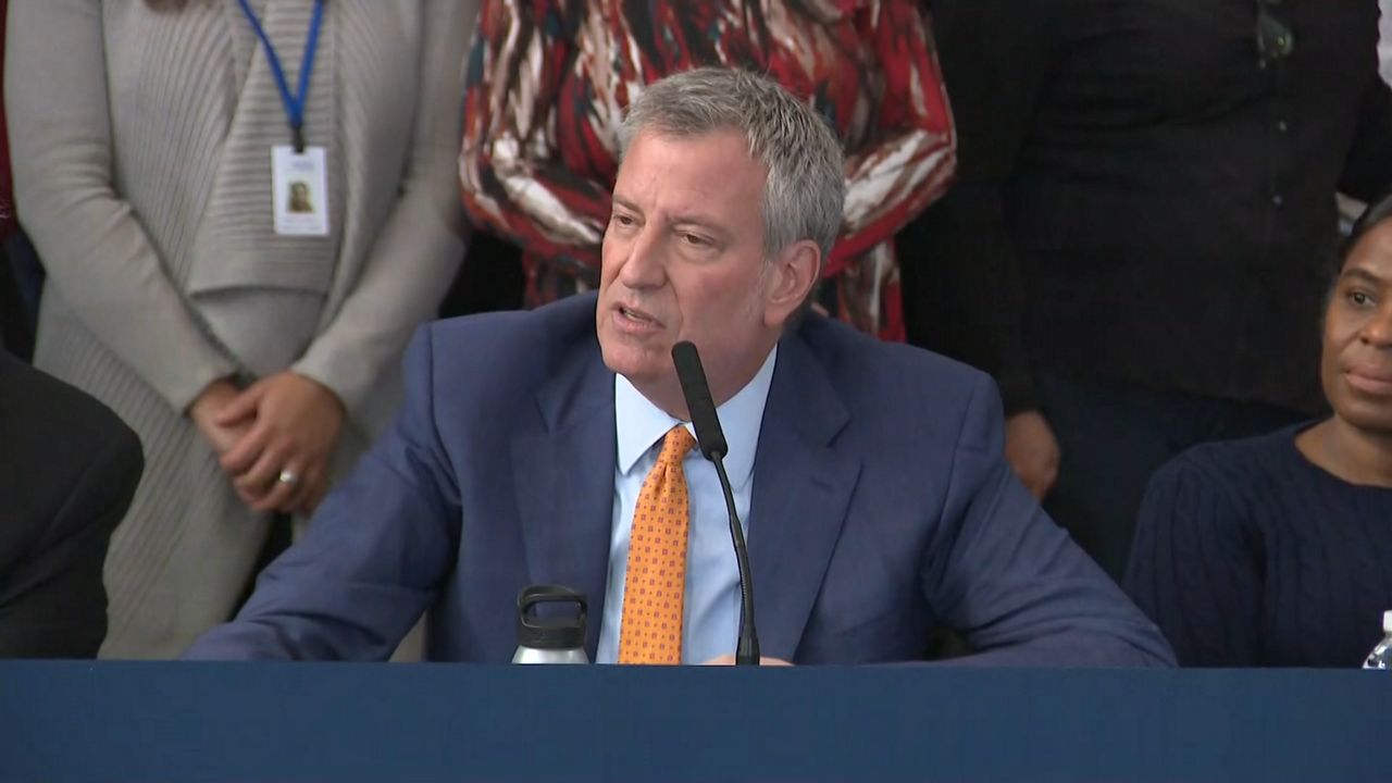 New York City Mayor Bill de Blasio, center, wearing a navy blue suit jacket, a sky blue dress shirt, a yellow tie with black spots, and speaking into a black microphone.
