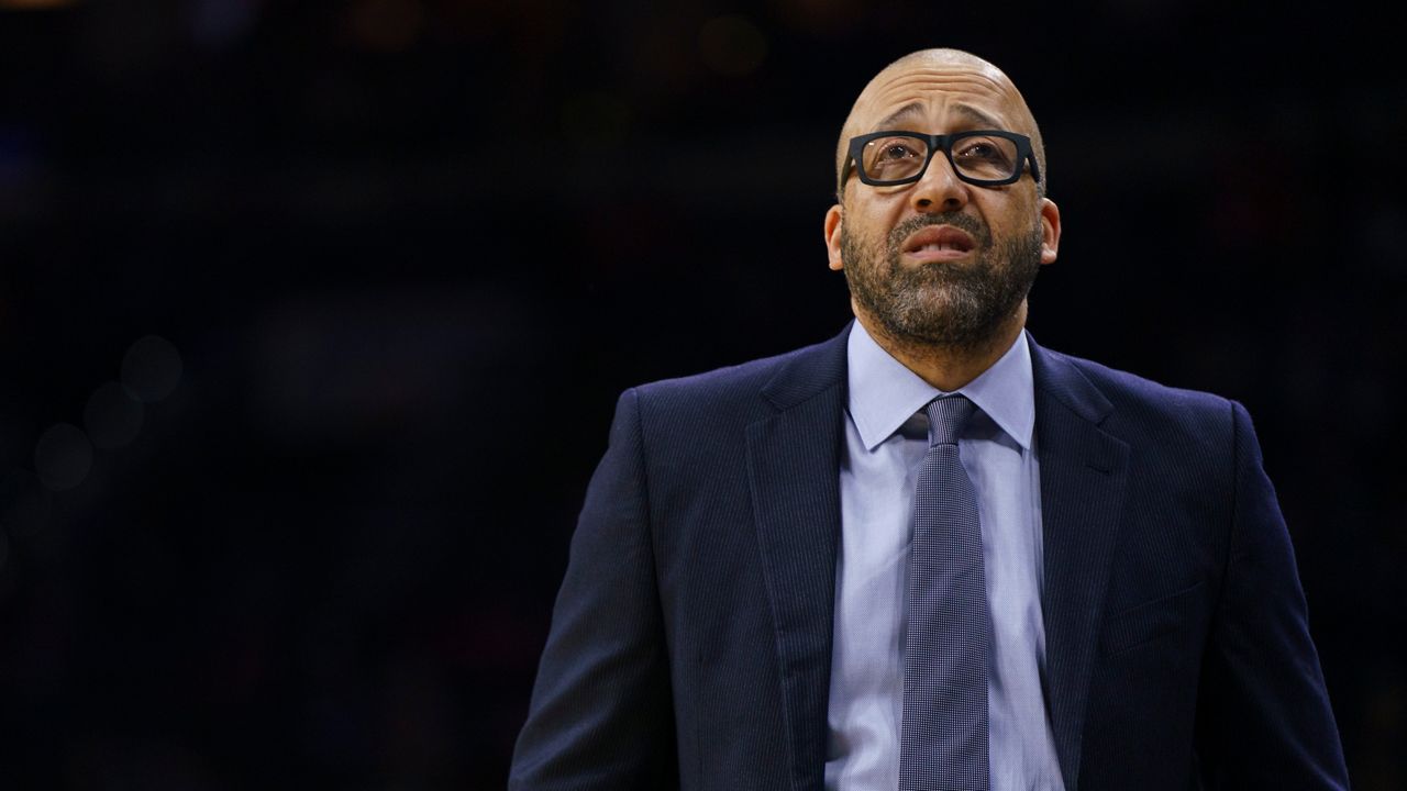 A man, wearing black, square-framed glasses, a navy blue suit jacket, a sky blue dress shirt, and a grey tie, stands in front of a solid black background.