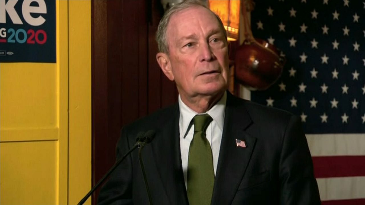 A man, wearing a black suit jacket, a white dress shirt, and a green tie, stands near a thin black microphone. A red-white-and-blue American flag hangs in the background.