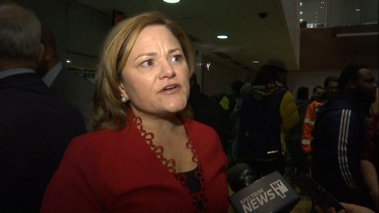 Melissa Mark-Viverito, wearing a black blouse and a red sweater, standing near a Spectrum News NY1 microphone.