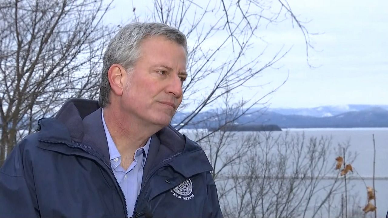 New York City Mayor Bill de Blasio, wearing a sky blue dress shirt and a navy blue coat, sits in front of a thicket of leafless tree branches near the sea.