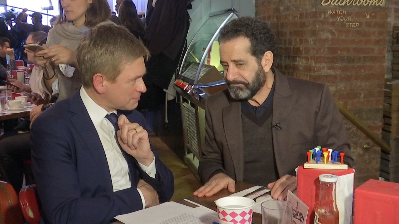 NY1 Anchor Pat Kiernan, left, wearing a white dress shirt and a navy blue tie and suit jacket. Tony Shalhoub, right, wearing a brown shirt and blazer. They sit at a brown table, with white papers, white cups with pink diamond patterns, and ketchup and white napkins in red containers. A red-brick pillar is behind them.