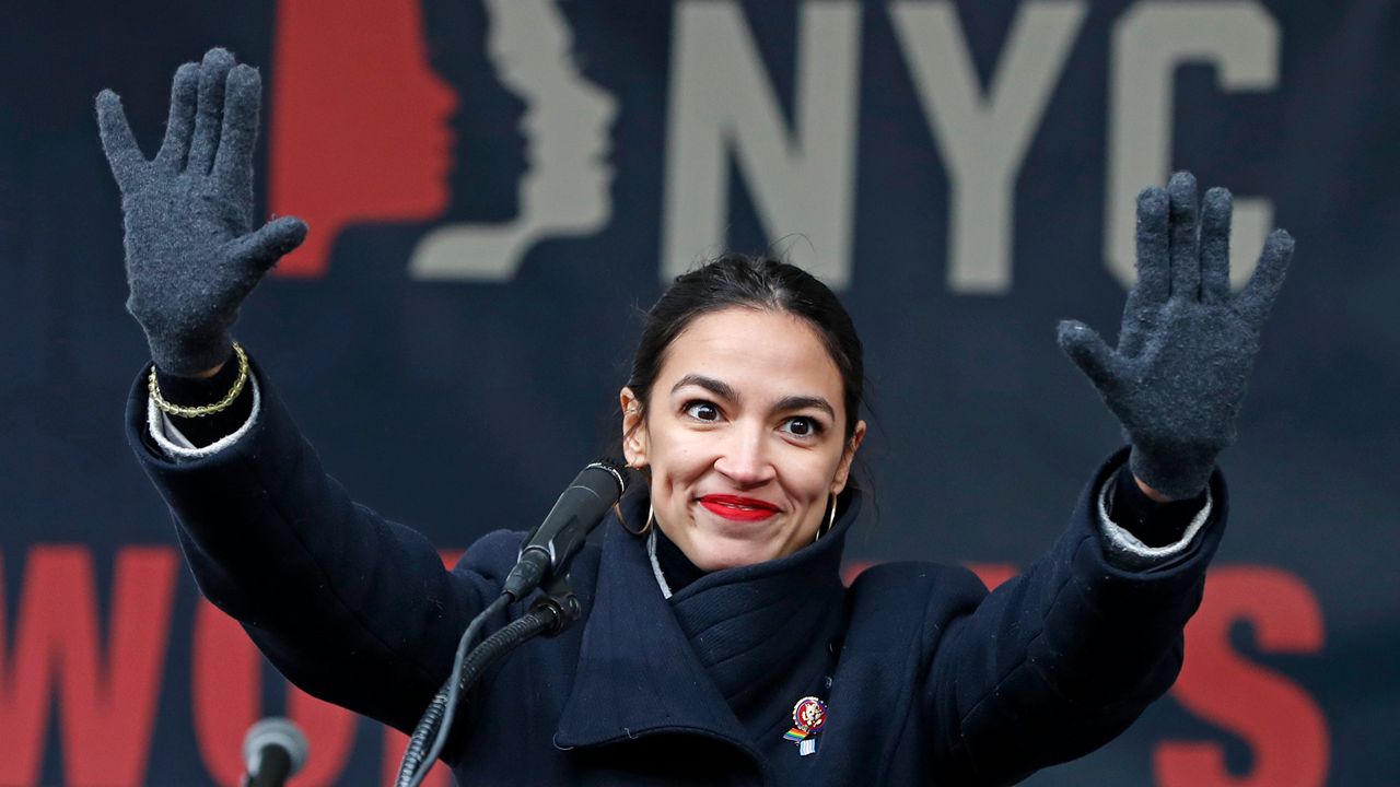 Alexandria Ocasio-Cortez, wearing dark blue gloves and a dark blue coat, stands near a black microphone in front of blue wallpaper.