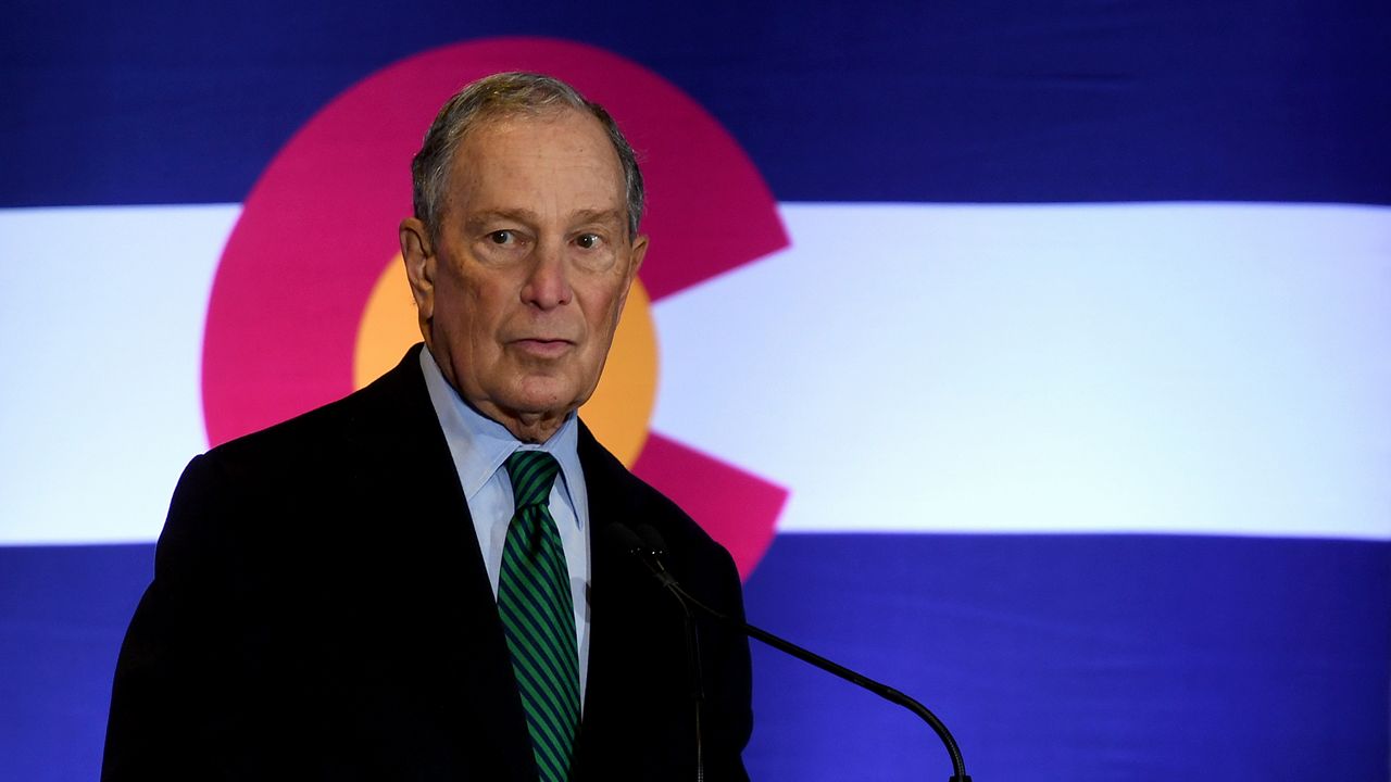 Michael Bloomberg, wearing a black suit jacket, a sky blue dress shirt, and a green tie with diagonal black stripes, speaks into a thin, black microphone near his chest. A blue-and-white Colorado state flag, with a Violet "C' enclosing a yellow sphere, is behind him.