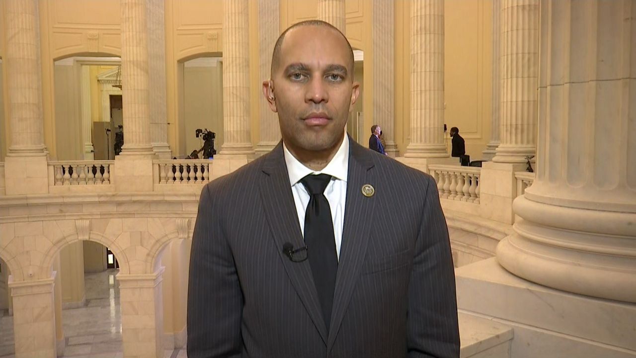 A man, wearing a black suit jacket, a black tie, and a white dress shirt, stands near a beige marble pillar that stands to his left in a great hall.