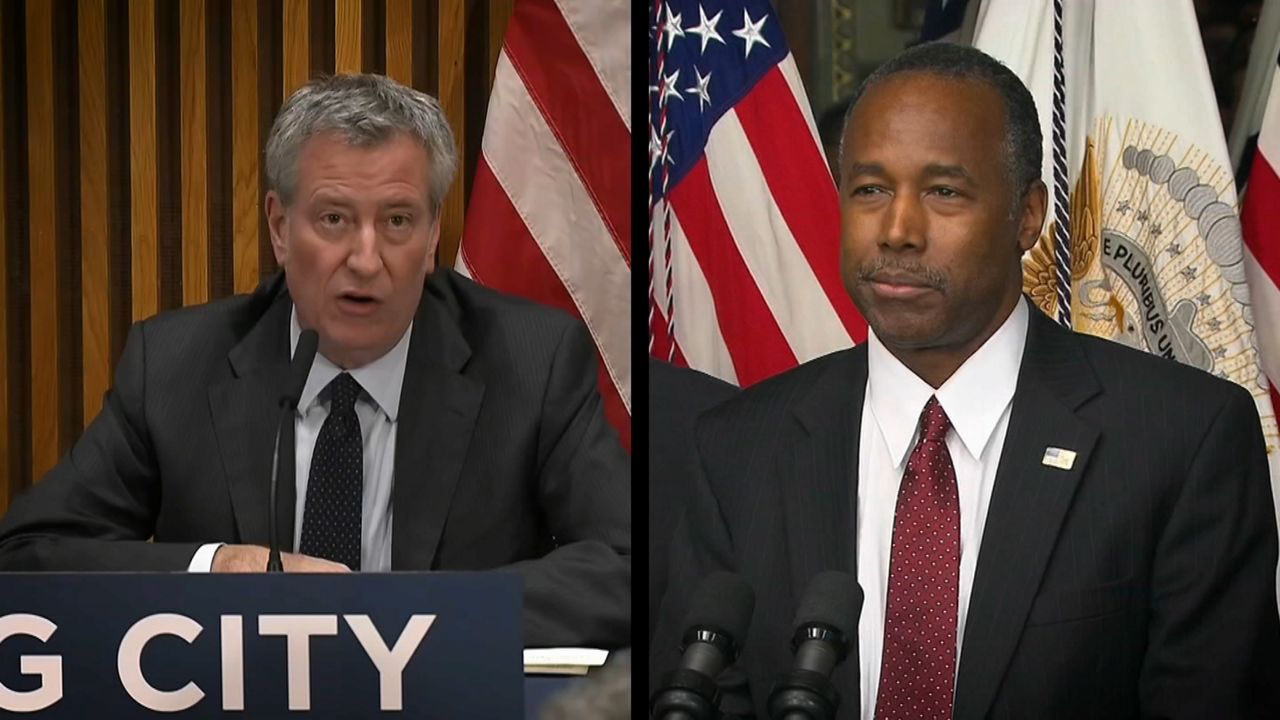 From left to right: Mayor Bill de Blasio, wearing a black suit jacket, a white dress shirt, and a black tie, sitting in front of a black microphone with an American flag over his left shoulder; HUD Secretary Ben Carson, wearing a black suit jacket, a white dress shirt, and a red tie with white spots, stands in front of two black microphones with an American flag behind his right shoulder.