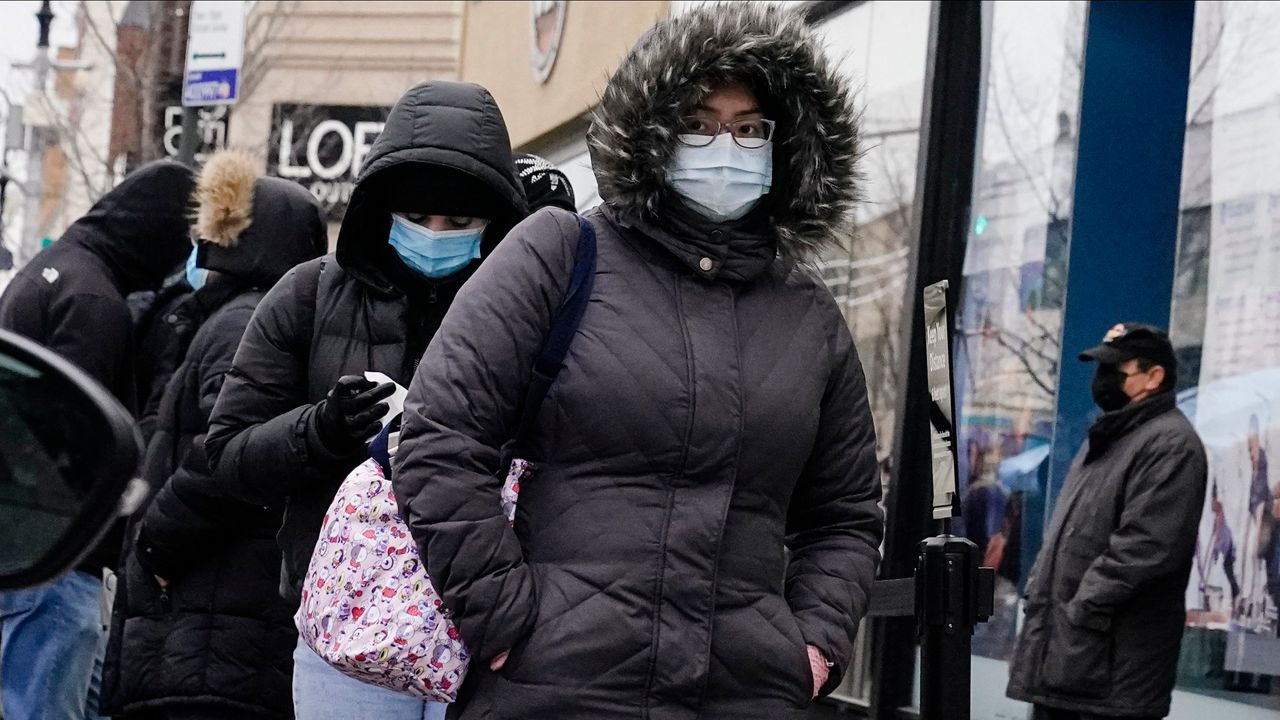A person, center, wearing a black jacket with a fur-lined hood, wears a sky blue medical mask and black, rectangular glasses, while waiting on line for COVID-19 testing at a CityMD in Forest Hills, Queens