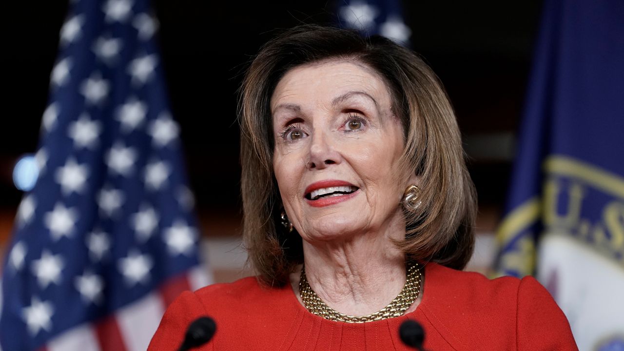 House Speaker Nancy Pelosi, wearing a red blouse, earrings, and a gold-colored necklace, speaks into two thin, black microphones, while standing in front of a red-white-and-blue American flag behind her right shoulder and a blue flag behind her left shoulder.