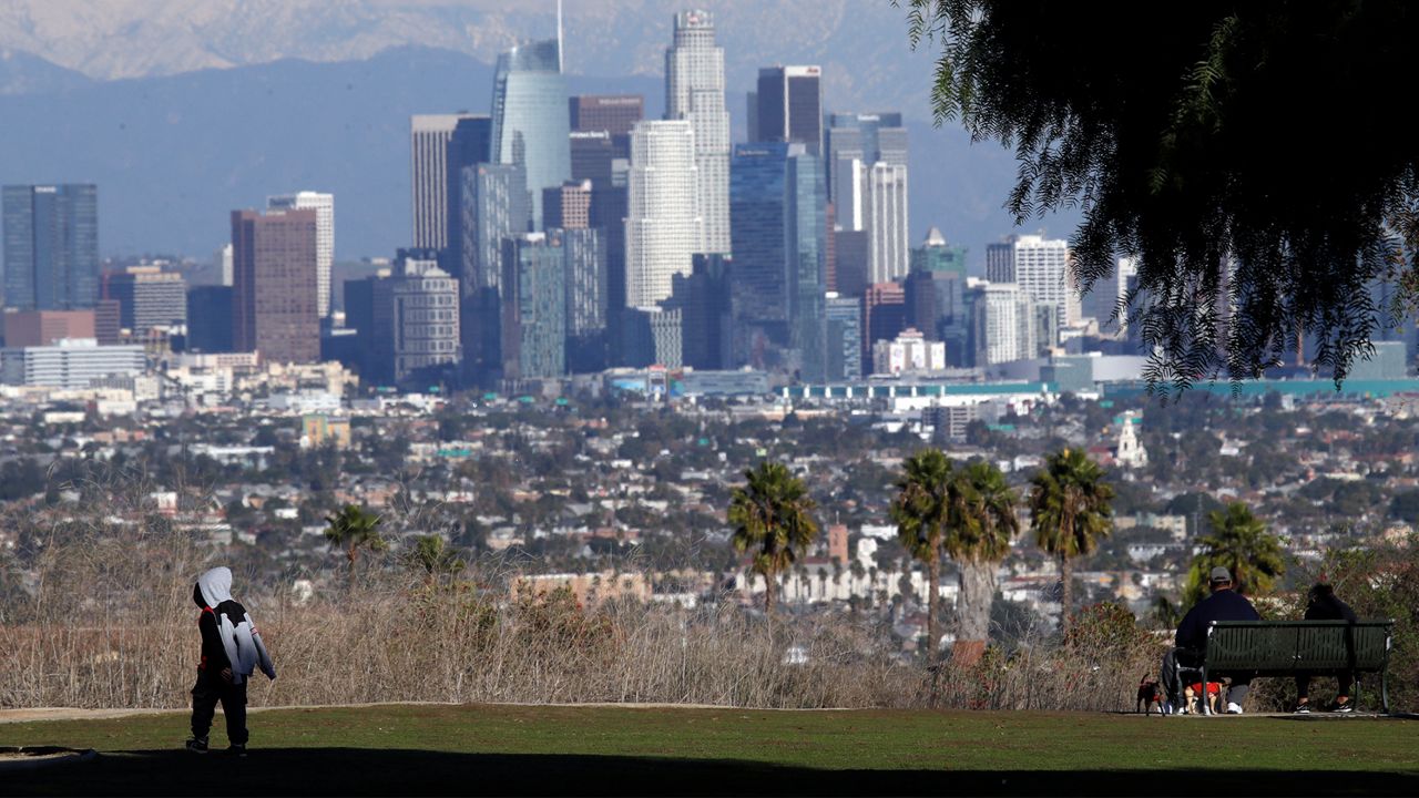 A boys walks under the shade from a tree in front of the snow-capped San Gabriel Mountains and the downtown skyline after a series of storms Friday, Dec. 27, 2019, in Los Angeles. (AP Photo/Marcio Jose Sanchez)