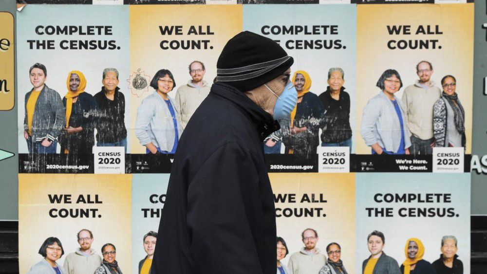 In this April 1, 2020, file photo, a man wearing a mask walks past posters encouraging participation in the 2020 Census in Seattle's Capitol Hill neighborhood. A delay in census data is scrambling plans in some states to redraw districts for the U.S. House and state legislatures. (AP Photo/Ted S. Warren, File)