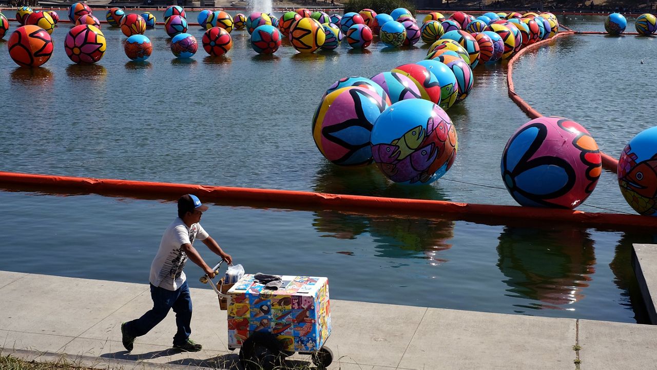 In this Aug. 22, 2015, file photo, an ice-cream vendor walks lakeside as spheres are being lowered into Los Angeles’ MacArthur Park Lake as part of a public art installation by Portraits of Hope. (AP Photo/Richard Vogel)