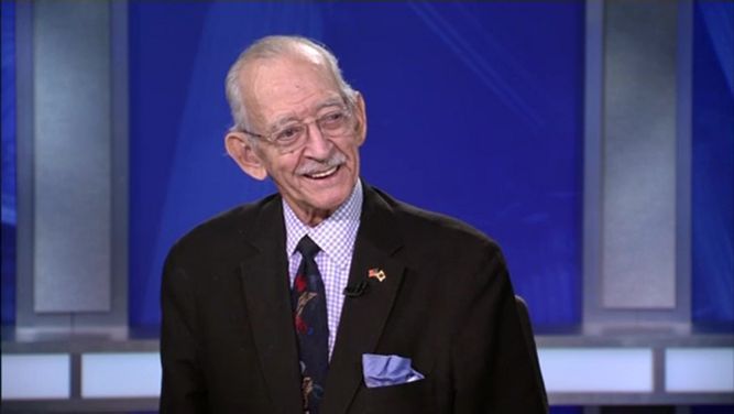 A man wearing square glasses, a black suit jacket, a plaid dress shirt, and a black tie sits on the set of NY1 political show "Road to City Hall."