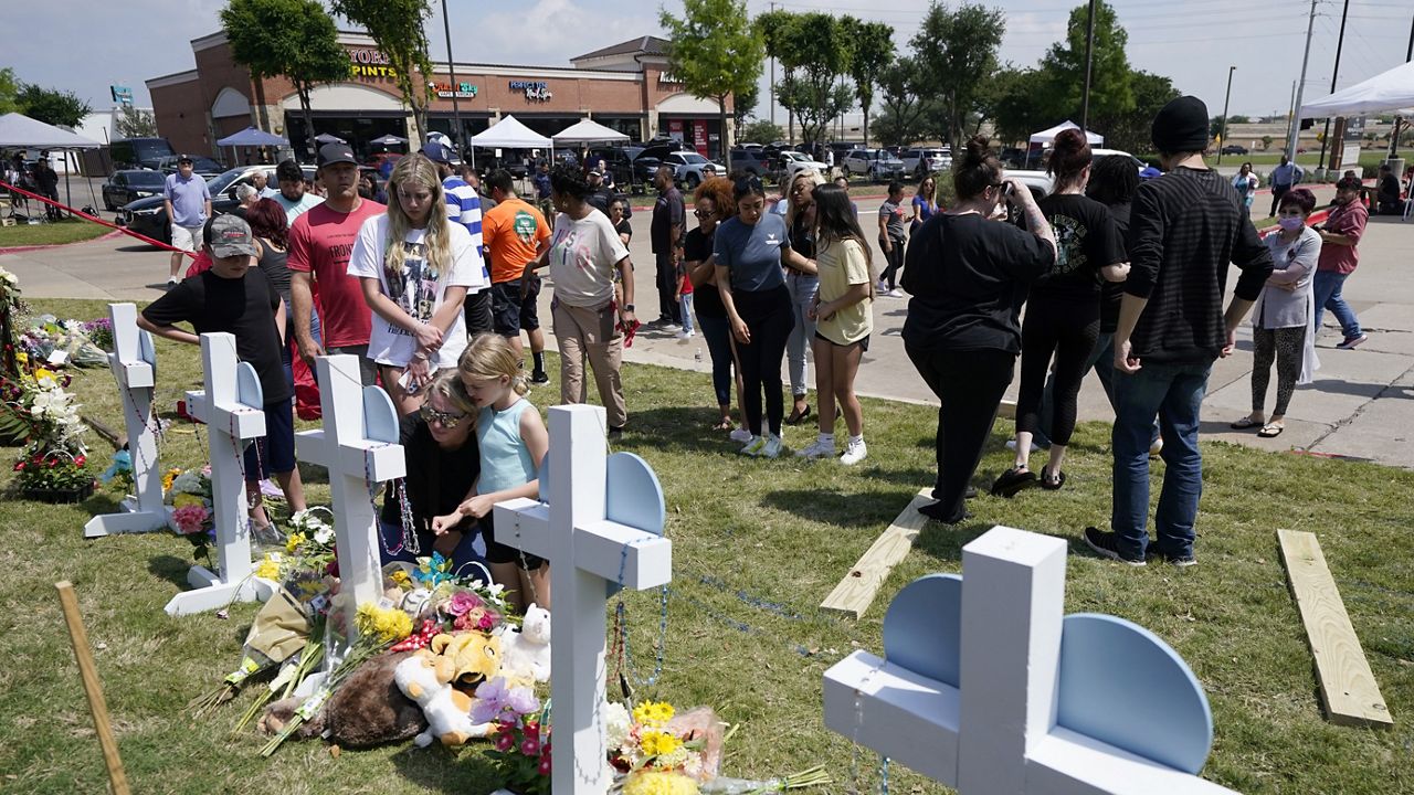 Jssica Himes, kneeling in front of cross, holds her daughter Harper, blue shirt, as her son Hudson, left in black shirt, her husband Scott, left rear in red shirt, and daughter Blakely Brooks, center left in white shirt, all from Allen, Texas, look on at a makeshift memorial by the mall where several people were killed in Saturday's mass shooting, Monday, May 8, 2023, in Allen, Texas. (AP Photo/Tony Gutierrez)