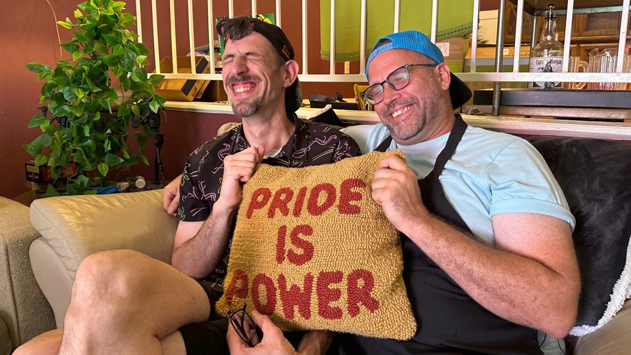 Couple Matthew Kistler and Daniel Cates smile inside their Fort Worth home with a couch pillow that reads "Pride is Power." (Spectrum News 1/Lupe Zapata)