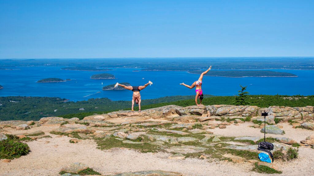 A man and a woman in athletic clothing do handstand poses in front of a camera on a tripod they have set up at the top of Cadillac Mountain in Acadia National Park, Maine, on Monday, August 16, 2021. (AP Photo/Ted Shaffrey)