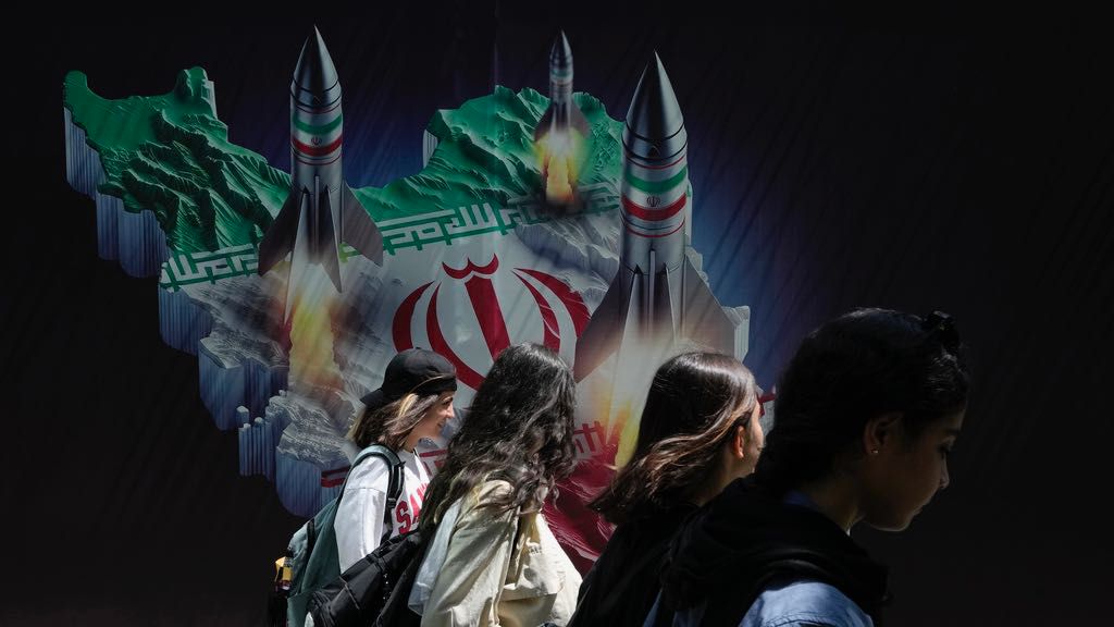 Iranian women without wearing their mandatory Islamic headscarf walk past a banner showing missiles being launched from Iranian map in northern Tehran, Iran, Friday, April 19, 2024. (AP Photo/Vahid Salemi)